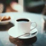 Coffee Loophole Recipe – Energizing Weight Loss Coffee That Works Fast 9 Coffee loophole recipe served in a white cup with black coffee on a saucer, with a spoon and soft background treats on a reflective table.