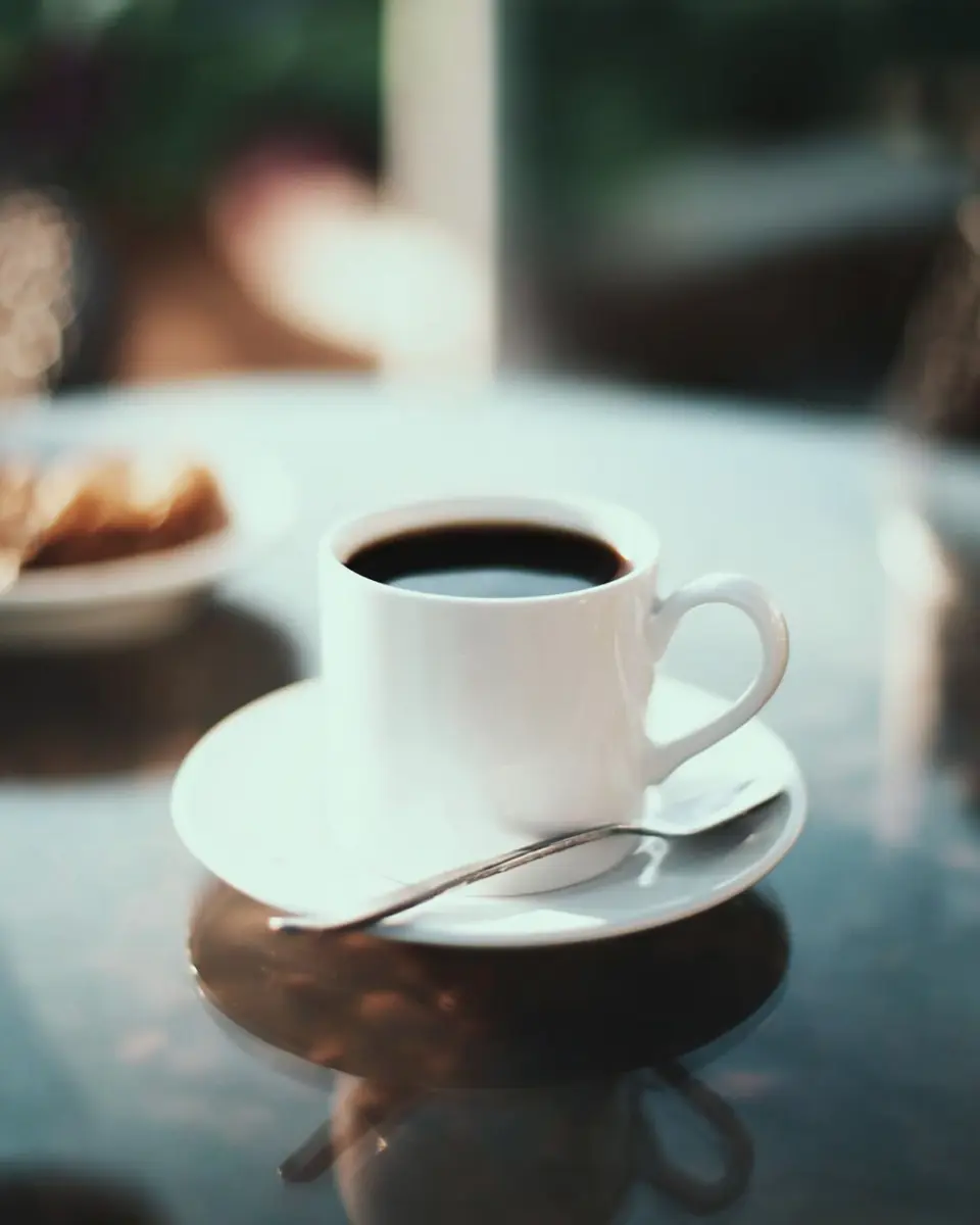 Coffee loophole recipe served in a white cup with black coffee on a saucer, with a spoon and soft background treats on a reflective table.