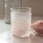 Close-up of a simple glass prepared for the pink salt trick recipe, filled with sole water and placed beside a bowl of pink Himalayan salt on a countertop