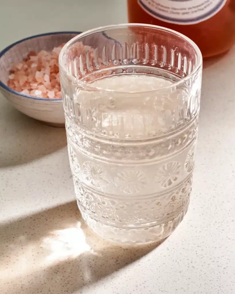 Glass of pink salt trick recipe water on a kitchen counter with pink salt and apple cider vinegar in the background