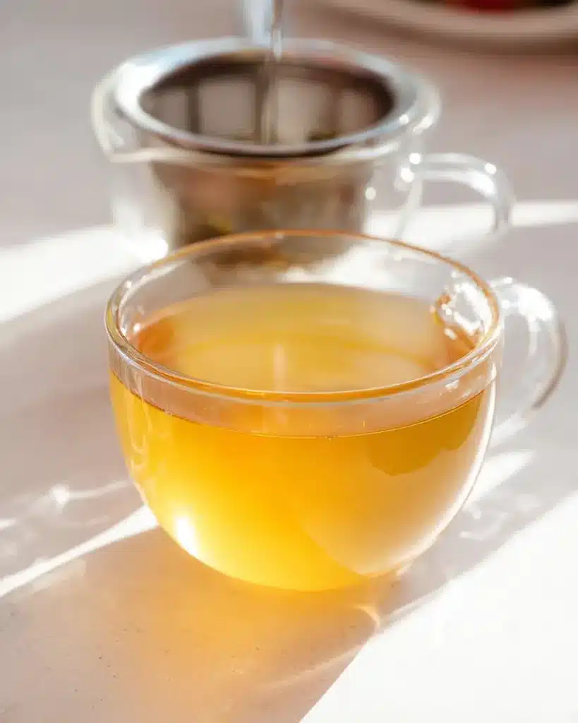 Tummy Tea Recipe for Belly Bloat, Digestion & Weight Loss Support 11 Golden tummy tea being poured into a glass cup with a tea strainer in the background