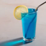 Blue tonic drink in a clear glass with lemon slice and metal straw on a light surface