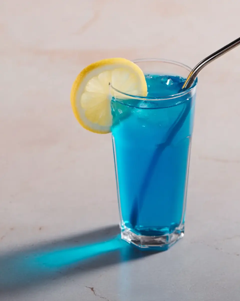 Blue tonic drink in a clear glass with lemon slice and metal straw on a light surface