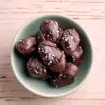 How to Make the Best Tahini Chocolate Date Dessert (Easy, Healthy & Irresistible) 7 Chocolate-covered dates topped with flaky sea salt in a pastel bowl on a wooden table