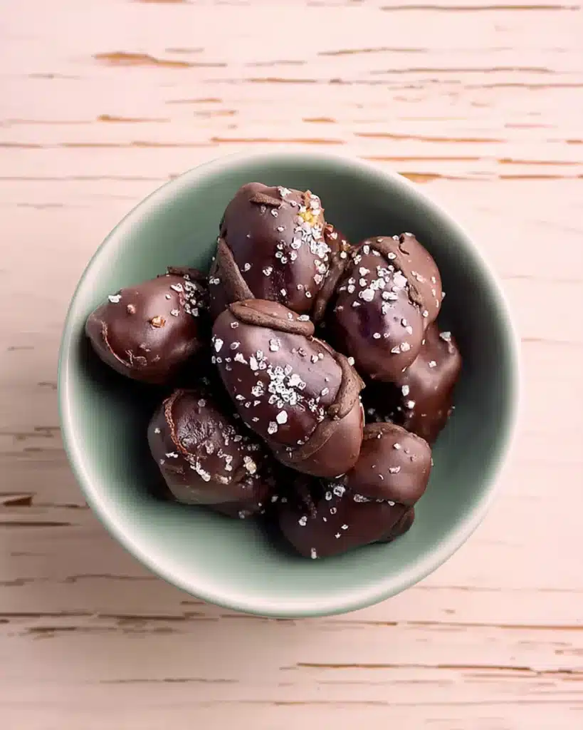 Chocolate-covered dates topped with flaky sea salt in a pastel bowl on a wooden table