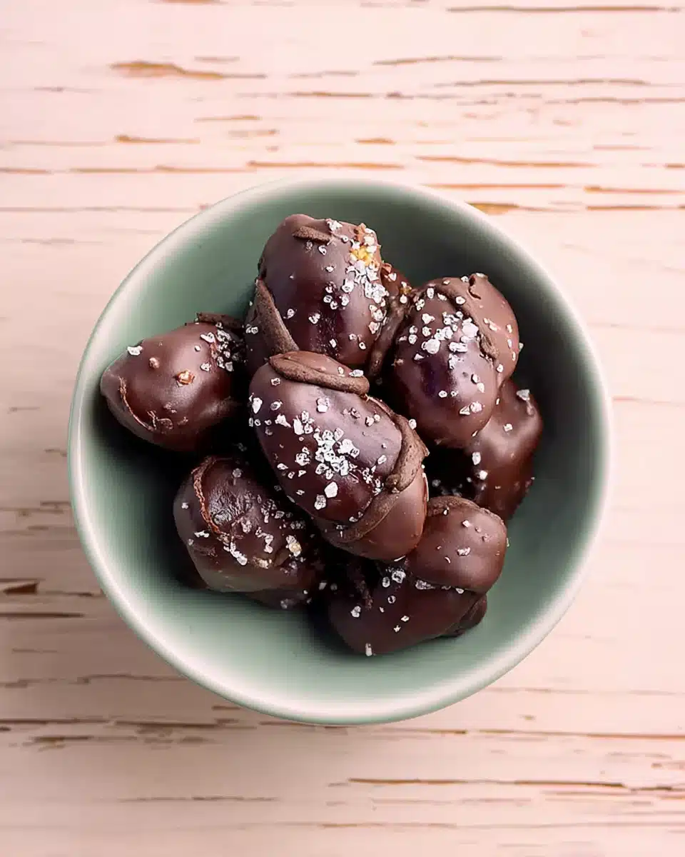 Chocolate-covered dates topped with flaky sea salt in a pastel bowl on a wooden table