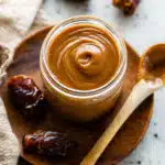 Dates Caramel Sauce Recipe – A Healthy, 3-Ingredient Natural Sweetener 7 Overhead view of a glass jar filled with caramel from a dates caramel sauce recipe, placed on a wooden plate with a spoon and surrounded by dried dates.