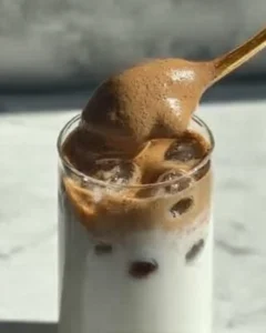 Spoon placing whipped honey coffee recipe foam over iced milk in a tall glass with visible ice cubes