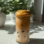 Whipped Honey Coffee Recipe: A Creamy, Naturally Sweet Dalgona Twist 17 Tall glass of whipped honey coffee with ice and frothy foam, sitting on a stone counter with plant in background