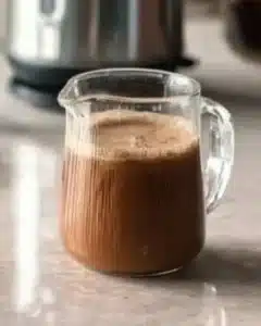 Glass jug with whipped honey coffee recipe base before mixing, filled with hot water, honey and instant coffee on marble counter