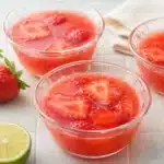 Strawberry Gelatin in glass bowls with sliced strawberries and lime on a bright surface