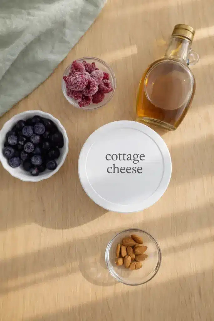 Cottage Cheese Breakfast Bowl for a 5 Minute High Protein Breakfast 9 Cottage Cheese Breakfast Bowl ingredients with berries, almonds, and maple syrup on a countertop
